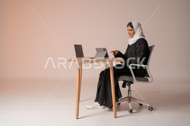 Using modern and advanced technologies, portrait of a veiled Saudi Arabian Gulf woman smiling wearing a black abaya sitting on a chair in front of a laptop smiling and pointing her index finger at the tablet making a video call, communicating with family and friends remotely, beige background