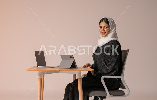 استخدام التقنيات الحديثة والمتطورة، بورتريه لامراة عربية خليجية سعودية محجبة مبتسمة ترتدي العباءة السوداء تجلس على الكرسي أمام الحاسوب المحمول مبتسمة تنظر الى الكميرا تقوم بإجراء مكالمة فيديو، التواصل مع الأهل و الأصدقاء عن بعد، خلفية بيج