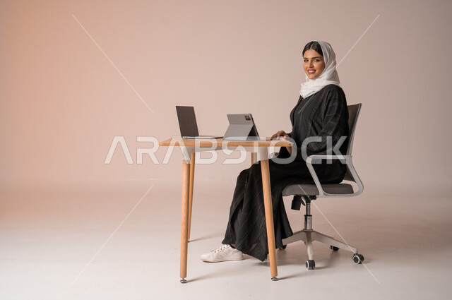 استخدام التقنيات الحديثة والمتطورة، بورتريه لامراة عربية خليجية سعودية محجبة مبتسمة ترتدي العباءة السوداء تجلس على الكرسي أمام الحاسوب المحمول مبتسمة تنظر الى الكميرا تقوم بإجراء مكالمة فيديو، التواصل مع الأهل و الأصدقاء عن بعد، خلفية بيج