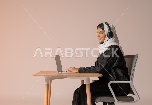 التواصل مع الأهل و الأصدقاء عن بعد، الرد على الاستعلامات والاستفسارات وخدمة العملاء، بورتريه لامراة عربية خليجية سعودية محجبة ترتدي العباءة السوداء مبتسمة تجلس على الكرسي امام الحاسوب المحمول وتضع سماعة الهيدفون، استخدام التقنيات الحديثة والمتطورة، خلفية بيج
