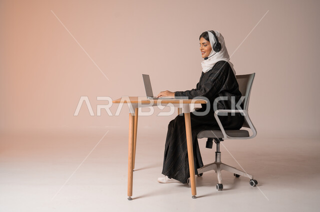 التواصل مع الأهل و الأصدقاء عن بعد، الرد على الاستعلامات والاستفسارات وخدمة العملاء، بورتريه لامراة عربية خليجية سعودية محجبة ترتدي العباءة السوداء مبتسمة تجلس على الكرسي امام الحاسوب المحمول وتضع سماعة الهيدفون، استخدام التقنيات الحديثة والمتطورة، خلفية بيج