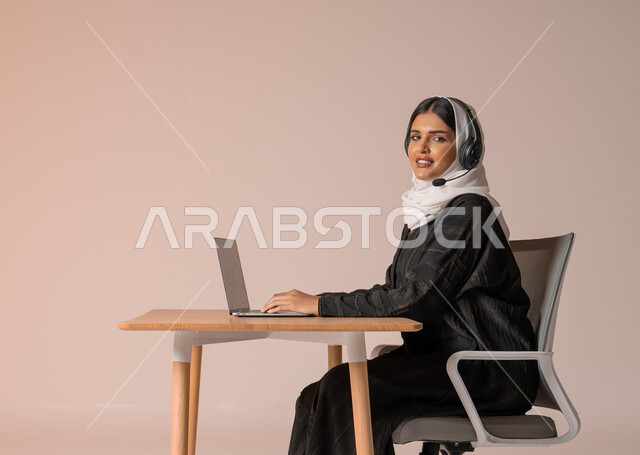 الرد على الاستعلامات والاستفسارات وخدمة العملاء، بورتريه لامراة عربية خليجية سعودية محجبة ترتدي العباءة السوداء مبتسمة تنظر الى الكاميرا تجلس على الكرسي امام الحاسوب المحمول وتضع سماعة الهيدفون، استخدام التقنيات الحديثة والمتطورة، التواصل مع الأهل و الأصدقاء عن بعد، خلفية بيج