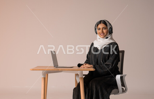 الرد على الاستعلامات والاستفسارات وخدمة العملاء، بورتريه لامراة عربية خليجية سعودية محجبة ترتدي العباءة السوداء مبتسمة تنظر الى الكاميرا تجلس على الكرسي امام الحاسوب المحمول وتضع سماعة الهيدفون، استخدام التقنيات الحديثة والمتطورة، التواصل مع الأهل و الأصدقاء عن بعد، خلفية بيج