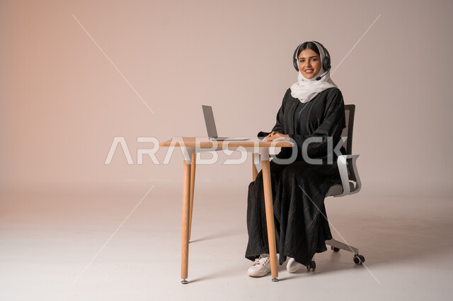 الرد على الاستعلامات والاستفسارات وخدمة العملاء، بورتريه لامراة عربية خليجية سعودية محجبة ترتدي العباءة السوداء مبتسمة تنظر الى الكاميرا تجلس على الكرسي امام الحاسوب المحمول وتضع سماعة الهيدفون، استخدام التقنيات الحديثة والمتطورة، التواصل مع الأهل و الأصدقاء عن بعد، خلفية بيج