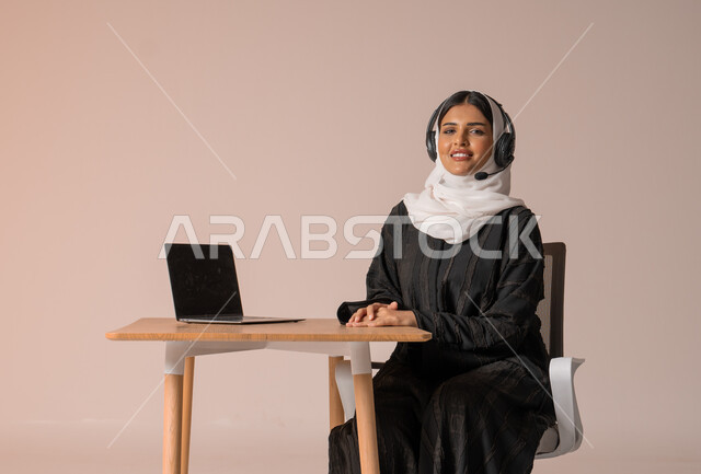 الرد على الاستعلامات والاستفسارات وخدمة العملاء، بورتريه لامراة عربية خليجية سعودية محجبة ترتدي العباءة السوداء مبتسمة تنظر الى الكاميرا تجلس على الكرسي امام الحاسوب المحمول وتضع سماعة الهيدفون، استخدام التقنيات الحديثة والمتطورة، التواصل مع الأهل و الأصدقاء عن بعد، خلفية بيج