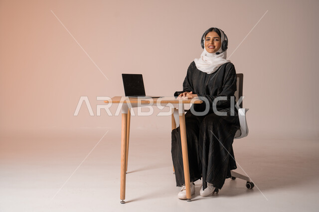 الرد على الاستعلامات والاستفسارات وخدمة العملاء، بورتريه لامراة عربية خليجية سعودية محجبة ترتدي العباءة السوداء مبتسمة تنظر الى الكاميرا تجلس على الكرسي امام الحاسوب المحمول وتضع سماعة الهيدفون، استخدام التقنيات الحديثة والمتطورة، التواصل مع الأهل و الأصدقاء عن بعد، خلفية بيج