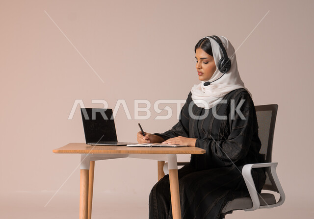 استخدام التقنيات الحديثة والمتطورة، الرد على الاستعلامات والاستفسارات وخدمة العملاء، بورتريه لامراة عربية خليجية سعودية محجبة ترتدي العباءة السوداء تجلس على الكرسي أمام الحاسوب المحمول وتضع سماعة الهيدفون، التواصل مع الأهل و الأصدقاء عن بعد، خلفية بيج