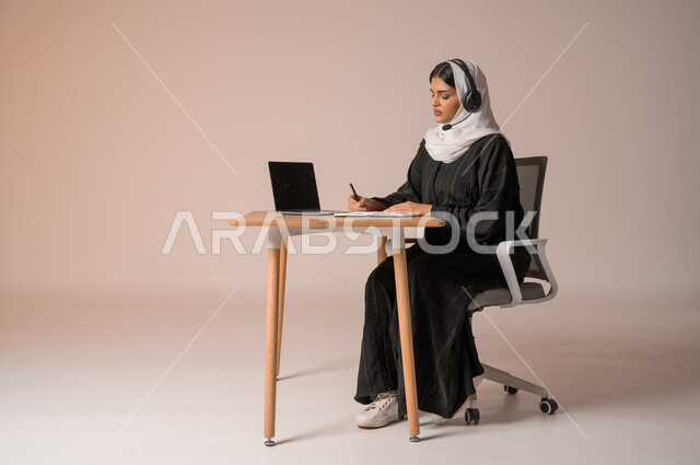 Using modern and advanced technologies, responding to inquiries and customer service, portrait of a veiled Saudi Arabian Gulf woman wearing a black abaya sitting on a chair in front of a laptop and wearing headphones, communicating with family and friends remotely, beige background