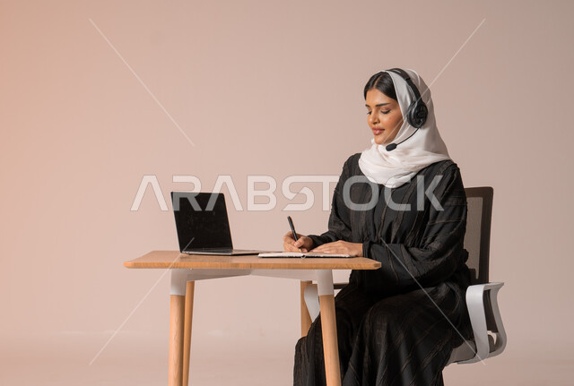 استخدام التقنيات الحديثة والمتطورة، الرد على الاستعلامات والاستفسارات وخدمة العملاء، بورتريه لامراة عربية خليجية سعودية محجبة ترتدي العباءة السوداء تجلس على الكرسي أمام الحاسوب المحمول وتضع سماعة الهيدفون، التواصل مع الأهل و الأصدقاء عن بعد، خلفية بيج