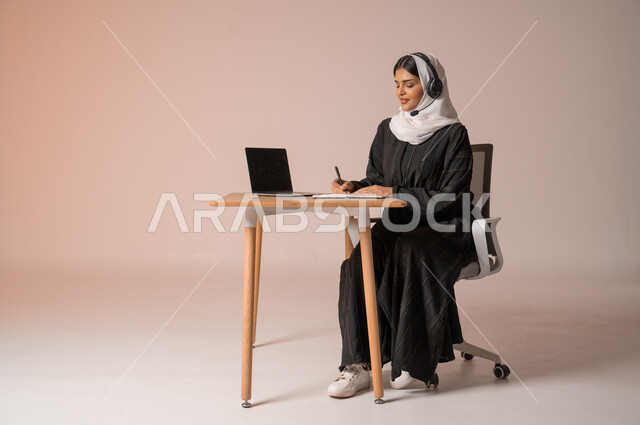 استخدام التقنيات الحديثة والمتطورة، الرد على الاستعلامات والاستفسارات وخدمة العملاء، بورتريه لامراة عربية خليجية سعودية محجبة ترتدي العباءة السوداء تجلس على الكرسي أمام الحاسوب المحمول وتضع سماعة الهيدفون، التواصل مع الأهل و الأصدقاء عن بعد، خلفية بيج