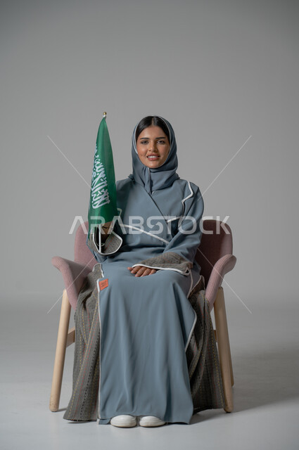  الذكرى والاحتفال بالعيد الوطني السعودي, بورتريه لامرأة عربية سعودية خليجية محجبة ترتدي عباءة تجلس على كرسي مريح تحمل بيدها علم المملكة العربية السعودية بايماءات السعادة والسرور، العلم السعودي راية التوحيد والإسلام, خلفية رمادية