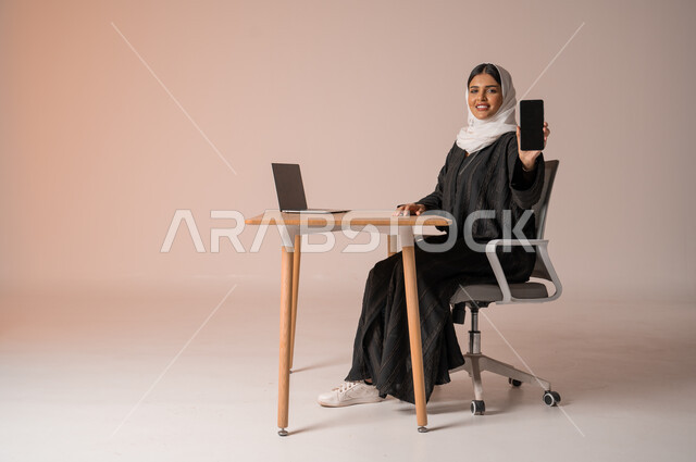 استخدام التقنيات الحديثة والمتطورة، بورتريه لامراة عربية خليجية سعودية محجبة مبتسمة ترتدي العباءة السوداء تجلس على الكرسي أمام الحاسوب المحمول مبتسمة تحمل بيدها الجوال بشاشة سوداء فارغه بإيماءات السرور، النظر الى الكاميرا بايماءات السعادة والسرور، خلفية بيج