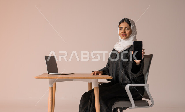 استخدام التقنيات الحديثة والمتطورة، بورتريه لامراة عربية خليجية سعودية محجبة مبتسمة ترتدي العباءة السوداء تجلس على الكرسي أمام الحاسوب المحمول مبتسمة تحمل بيدها الجوال بشاشة سوداء فارغه بإيماءات السرور، النظر الى الكاميرا بايماءات السعادة والسرور، خلفية بيج