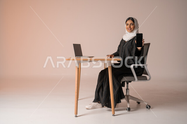 استخدام التقنيات الحديثة والمتطورة، بورتريه لامراة عربية خليجية سعودية محجبة مبتسمة ترتدي العباءة السوداء تجلس على الكرسي أمام الحاسوب المحمول مبتسمة تحمل بيدها الجوال بشاشة سوداء فارغه بإيماءات السرور، النظر الى الكاميرا بايماءات السعادة والسرور، خلفية بيج
