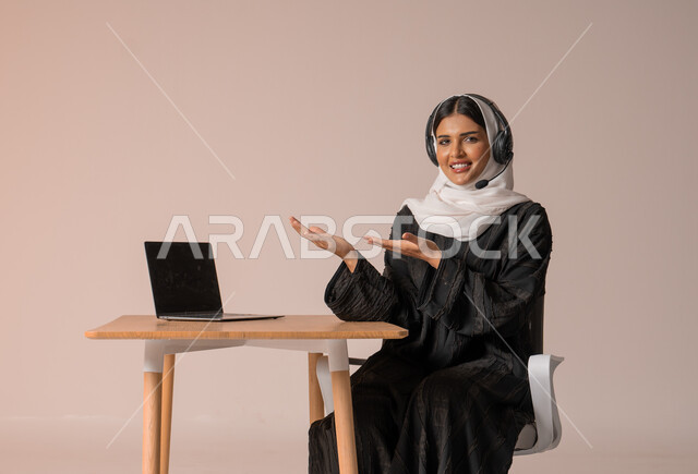 استخدام التقنيات الحديثة والمتطورة، الرد على الاستعلامات والاستفسارات وخدمة العملاء، بورتريه لامراة عربية خليجية سعودية محجبة ترتدي العباءة السوداء تجلس على الكرسي تشير بيديها الى الحاسوب المحمول وتضع سماعة الهيدفون، التواصل مع الأهل و الأصدقاء عن بعد، خلفية بيج