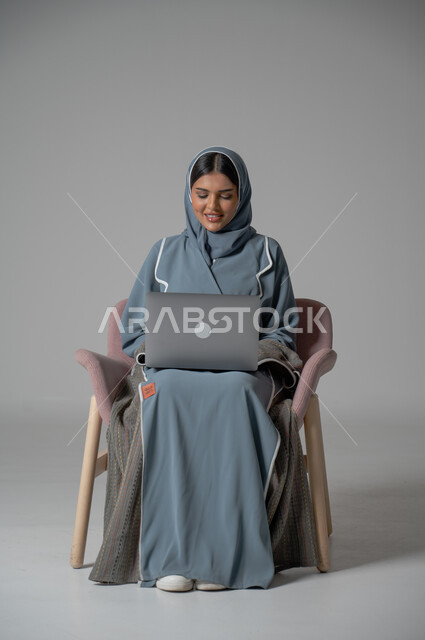 استخدام التقنيات الحديثة والمتطورة، بورتريه لامراة عربية خليجية سعودية محجبة مبتسمة ترتدي العباءة  تجلس على الكرسي تستخدم الحاسوب المحمول بايماءات السعادة والسرور، التواصل مع الأهل و الأصدقاء عن بعد، إدارة شؤون الأعمال وتنظيمه, خلفية رمادية