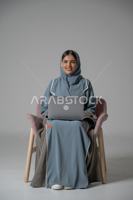 استخدام التقنيات الحديثة والمتطورة، بورتريه لامراة عربية خليجية سعودية محجبة مبتسمة ترتدي العباءة  تجلس على الكرسي تستخدم الحاسوب المحمول بايماءات السعادة والسرور، التواصل مع الأهل و الأصدقاء عن بعد، إدارة شؤون الأعمال وتنظيمه, خلفية رمادية