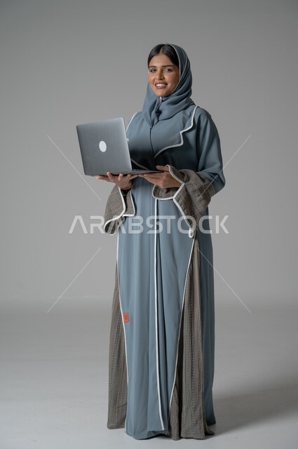  التواصل مع الأهل و الأصدقاء عن بعد، إدارة شؤون الأعمال وتنظيمه, استخدام التقنيات الحديثة والمتطورة، بورتريه لامراة عربية خليجية سعودية محجبة مبتسمة ترتدي العباءة تحمل بيدها الحاسوب المحمول بايماءات السعادة والسرور, صورة الجسم بالكامل, خلفية رمادية