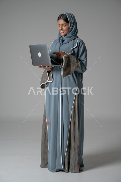 Communicating with family and friends remotely, managing and organizing business affairs, using modern and advanced technologies, portrait of a veiled Saudi Arabian Gulf woman, smiling, wearing an abaya, holding a laptop in her hand with gestures of happiness and pleasure, full body portrait, gray background