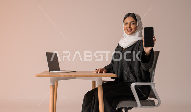 استخدام التقنيات الحديثة والمتطورة، بورتريه لامراة عربية خليجية سعودية محجبة مبتسمة ترتدي العباءة السوداء تجلس على الكرسي أمام الحاسوب المحمول مبتسمة تحمل بيدها الجوال بشاشة سوداء فارغه بإيماءات السرور، النظر الى الكاميرا بايماءات السعادة والسرور، خلفية بيج