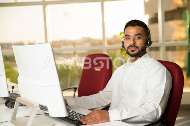 موظف عربي خليجي سعودي يعمل في خدمة العملاء ويرتدي سماعة الاتصال، استعلامات واستفسارات، الاجابه على اسئلة العملاء، اجراء اتصال فيديو من خلال الحاسوب، نظارة طبية، متابعة وتطوير العمل