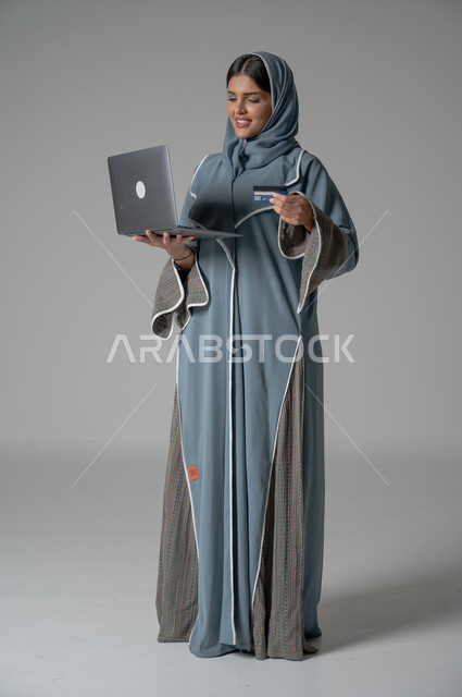 The possibility of purchasing and paying through electronic programs and applications, using modern technical devices for remote payment via the Internet, a portrait of a veiled Saudi Arabian Gulf woman wearing an abaya, holding a laptop and bank credit card in her hands for electronic shopping, full body portrait, gray background