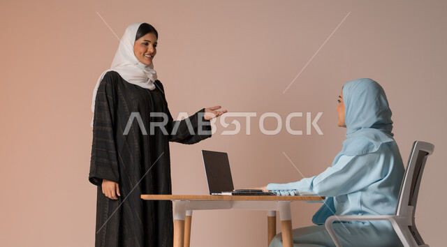 تبادل الخبرات والاتفاقيات الناجحة في مكتب الشركه، مفهوم الإدارة المتعاونة بين الموظفين، بورتريه لصديقتين عربيتين خليجيتين سعوديتين مبتسمتين ترتديان العباءة، استخدام جهاز الحاسوب المحمول لمتابعة ومناقشة الأعمال, خلفية بيج