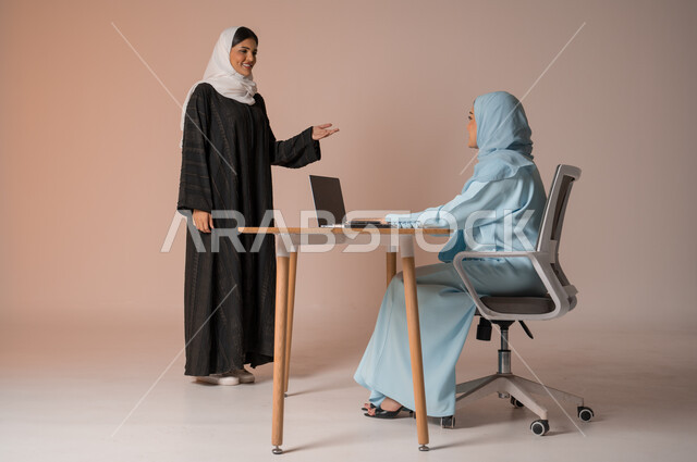 تبادل الخبرات والاتفاقيات الناجحة في مكتب الشركه، مفهوم الإدارة المتعاونة بين الموظفين، بورتريه لصديقتين عربيتين خليجيتين سعوديتين مبتسمتين ترتديان العباءة، استخدام جهاز الحاسوب المحمول لمتابعة ومناقشة الأعمال, خلفية بيج