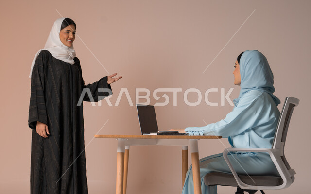 تبادل الخبرات والاتفاقيات الناجحة في مكتب الشركه، مفهوم الإدارة المتعاونة بين الموظفين، بورتريه لصديقتين عربيتين خليجيتين سعوديتين مبتسمتين ترتديان العباءة، استخدام جهاز الحاسوب المحمول لمتابعة ومناقشة الأعمال, خلفية بيج