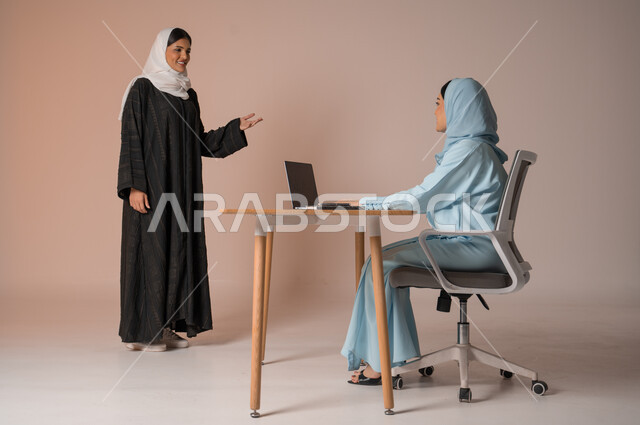 تبادل الخبرات والاتفاقيات الناجحة في مكتب الشركه، مفهوم الإدارة المتعاونة بين الموظفين، بورتريه لصديقتين عربيتين خليجيتين سعوديتين مبتسمتين ترتديان العباءة، استخدام جهاز الحاسوب المحمول لمتابعة ومناقشة الأعمال, خلفية بيج