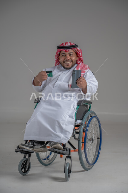 الشراء والدفع من خلال برامج وتطبيقات الجوال، بورتريه مقرب لرجل عربي خليجي سعودي مقعد قزم يرتدي الشماغ والثوب التقليدي يجلس على كرسي متحرك يحمل بيده الهاتف المحمل وبطاقة الإئتمان البنكية، تسديد الفواتير بالبطاقات الإلكترونية، خلفية رمادية