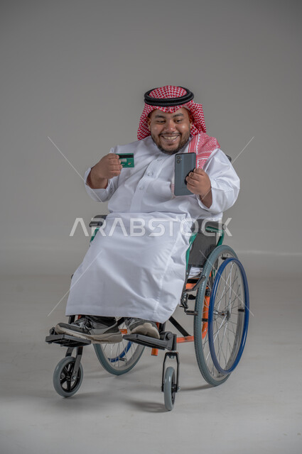 الشراء والدفع من خلال برامج وتطبيقات الجوال، بورتريه مقرب لرجل عربي خليجي سعودي مقعد قزم يرتدي الشماغ والثوب التقليدي يجلس على كرسي متحرك يحمل بيده الهاتف المحمل وبطاقة الإئتمان البنكية، تسديد الفواتير بالبطاقات الإلكترونية، خلفية رمادية