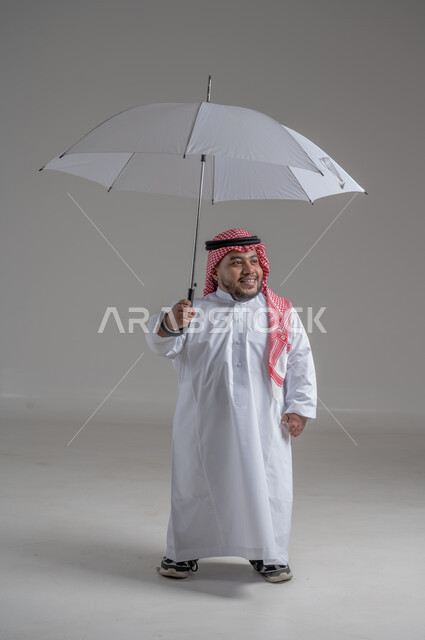 الحماية من الأمطار في الشتاء، تعابير البهجة والسرور، بورتريه لشاب عربي خليجي سعودي قزم مبتسم يرتدي الثوب التقليدي والشماغ يحمل بيده مظلة زرقاء، ايماءات الفرح والسعادة، التظلل من أشعة الشمس، صورة لكامل الجسم، خلفية رمادية