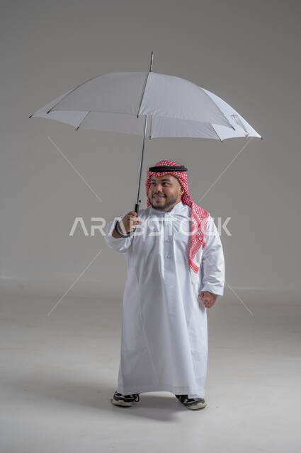 الحماية من الأمطار في الشتاء، تعابير البهجة والسرور، بورتريه لشاب عربي خليجي سعودي قزم مبتسم يرتدي الثوب التقليدي والشماغ يحمل بيده مظلة زرقاء، ايماءات الفرح والسعادة، التظلل من أشعة الشمس، صورة لكامل الجسم، خلفية رمادية