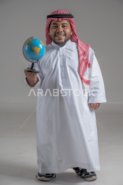 Locating Arab countries and states on the world map, searching and exploring the universe, portrait of a smiling Saudi Arabian Gulf dwarf man wearing traditional thobe and shemagh holding a globe in his hand, concept of travel and tours, full-length body, gray background