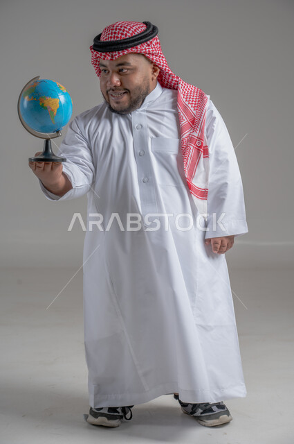 تحديد موقع الدول والبلدان العربية على خريطة العالم, البحث والاستكشاف حول الكون, بورتريه لرجل عربي خليجي سعودي قزم مبتسم يرتدي الثوب التقليدي والشماغ يحمل بيده مجسم الكرة الارضية, مفهوم الرحلات والجولات السياحية, الجسم بالطول الكامل، خلفية رمادية