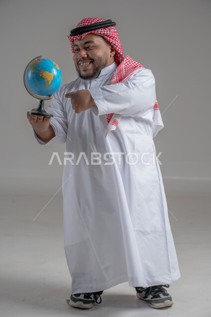 تحديد موقع الدول والبلدان العربية على خريطة العالم, البحث والاستكشاف حول الكون, بورتريه لرجل عربي خليجي سعودي قزم مبتسم يرتدي الثوب التقليدي والشماغ يحمل بيده مجسم الكرة الارضية, مفهوم الرحلات والجولات السياحية, الجسم بالطول الكامل، خلفية رمادية