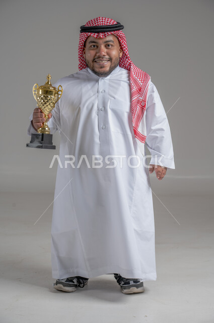مفهوم الفوز والانتصار، التتويج بالبطولة بعد النجاح، ايماءات السعادة والسرور، بورتريه لشاب عربي خليجي سعودي قزم يرتدي الثوب التقليدي والشماغ يقف باستقامة ويحمل بيده كأس الفوز، صورة الجسم بالطول الكامل، خلفية رمادية