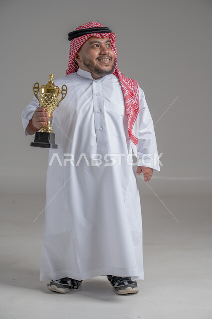 مفهوم الفوز والانتصار، التتويج بالبطولة بعد النجاح، ايماءات السعادة والسرور، بورتريه لشاب عربي خليجي سعودي قزم يرتدي الثوب التقليدي والشماغ يقف باستقامة ويحمل بيده كأس الفوز، صورة الجسم بالطول الكامل، خلفية رمادية