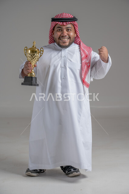 مفهوم الفوز والانتصار، التتويج بالبطولة بعد النجاح، ايماءات السعادة والسرور، بورتريه لشاب عربي خليجي سعودي قزم يرتدي الثوب التقليدي والشماغ يقف باستقامة ويحمل بيده كأس الفوز، صورة الجسم بالطول الكامل، خلفية رمادية