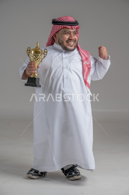 مفهوم الفوز والانتصار، التتويج بالبطولة بعد النجاح، ايماءات السعادة والسرور، بورتريه لشاب عربي خليجي سعودي قزم يرتدي الثوب التقليدي والشماغ يقف باستقامة ويحمل بيده كأس الفوز، صورة الجسم بالطول الكامل، خلفية رمادية
