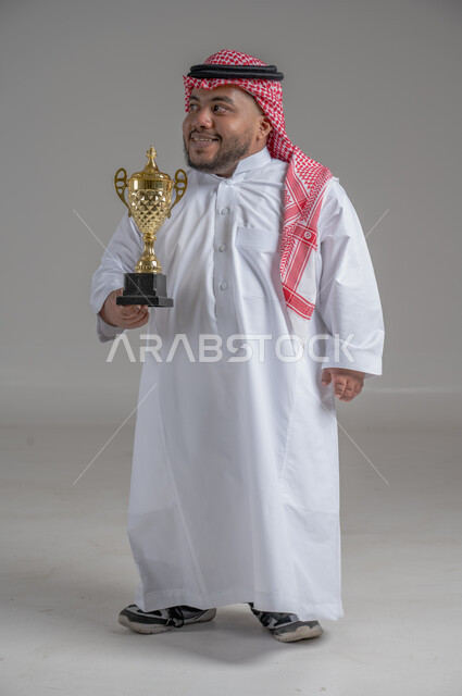 مفهوم الفوز والانتصار، التتويج بالبطولة بعد النجاح، ايماءات السعادة والسرور، بورتريه لشاب عربي خليجي سعودي قزم يرتدي الثوب التقليدي والشماغ يقف باستقامة ويحمل بيده كأس الفوز، صورة الجسم بالطول الكامل، خلفية رمادية