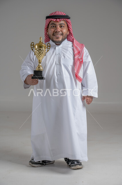 مفهوم الفوز والانتصار، التتويج بالبطولة بعد النجاح، ايماءات السعادة والسرور، بورتريه لشاب عربي خليجي سعودي قزم يرتدي الثوب التقليدي والشماغ يقف باستقامة ويحمل بيده كأس الفوز، صورة الجسم بالطول الكامل، خلفية رمادية