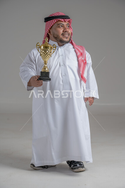 مفهوم الفوز والانتصار، التتويج بالبطولة بعد النجاح، ايماءات السعادة والسرور، بورتريه لشاب عربي خليجي سعودي قزم يرتدي الثوب التقليدي والشماغ يقف باستقامة ويحمل بيده كأس الفوز، صورة الجسم بالطول الكامل، خلفية رمادية