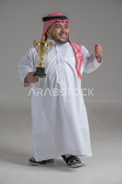 مفهوم الفوز والانتصار، التتويج بالبطولة بعد النجاح، ايماءات السعادة والسرور، بورتريه لشاب عربي خليجي سعودي قزم يرتدي الثوب التقليدي والشماغ يقف باستقامة ويحمل بيده كأس الفوز، صورة الجسم بالطول الكامل، خلفية رمادية