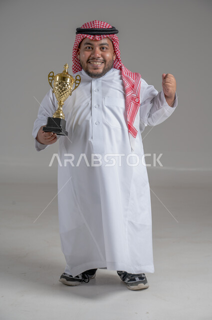 مفهوم الفوز والانتصار، التتويج بالبطولة بعد النجاح، ايماءات السعادة والسرور، بورتريه لشاب عربي خليجي سعودي قزم يرتدي الثوب التقليدي والشماغ يقف باستقامة ويحمل بيده كأس الفوز، صورة الجسم بالطول الكامل، خلفية رمادية