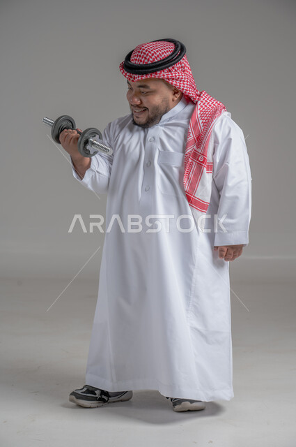 تمرين رفع الأثقال، القوة والتحدي والتحفيز لأسلوب حياة صحي، نشاط بدني للمحافظة على الصحة واللياقة الجسدية، رفع الأوزان الحديدية الثقيلة، تقوية عضلات الجسم وبناؤها، بورتريه مقرب لشاب عربي خليجي سعودي قزم يمارس التمارين الرياضية مع الدمبلز، خلفية رمادية