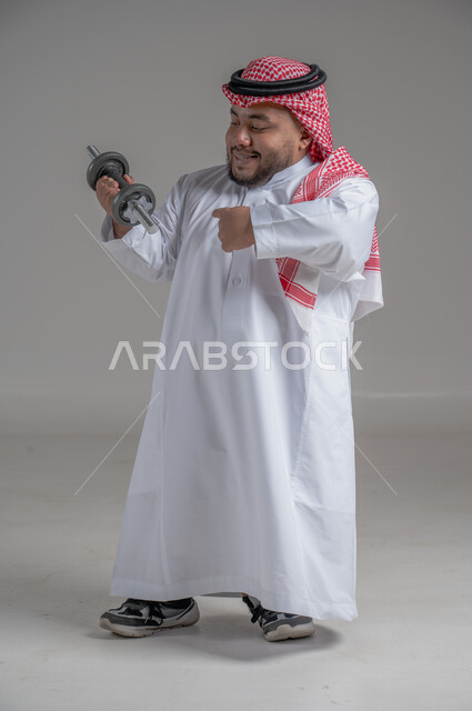 تمرين رفع الأثقال، القوة والتحدي والتحفيز لأسلوب حياة صحي، نشاط بدني للمحافظة على الصحة واللياقة الجسدية، رفع الأوزان الحديدية الثقيلة، تقوية عضلات الجسم وبناؤها، بورتريه مقرب لشاب عربي خليجي سعودي قزم يمارس التمارين الرياضية مع الدمبلز، خلفية رمادية