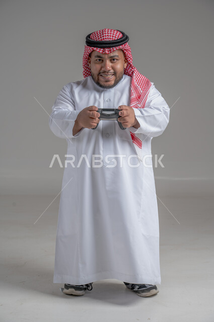 Spending free time playing PlayStation entertainment games, gestures of happiness and pleasure, entertaining modern technologies, portrait of a young Saudi Gulf dwarf with special needs wearing traditional thobe and shemagh holding a joystick in his hand, full-length body image, gray background