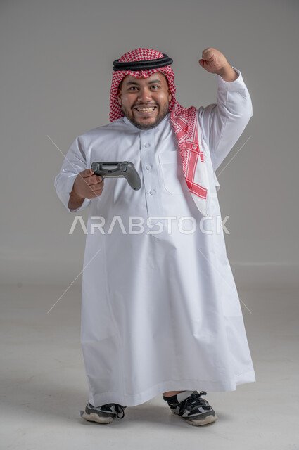 قضاء أوقات الفراغ باللعب بألعاب البلايستيشن الترفيهية ، إيماءات السعادة والسرور, تقنيات حديثة مسلية, بورتريه لشاب سعودي خليجي قزم  من ذوي الاحتياجات الخاصة يرتدي الثوب التقليدي والشماغ  يحمل بيده عصا التحكم، صورة الجسم بالطول الكامل، خلفية رمادية